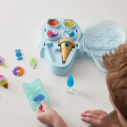 Child arranging blue and purple Aquabeads in a small turquoise tray, with the open ice cream storage case, ice cream bead pen, water sprayer and finished bead creations on the table.