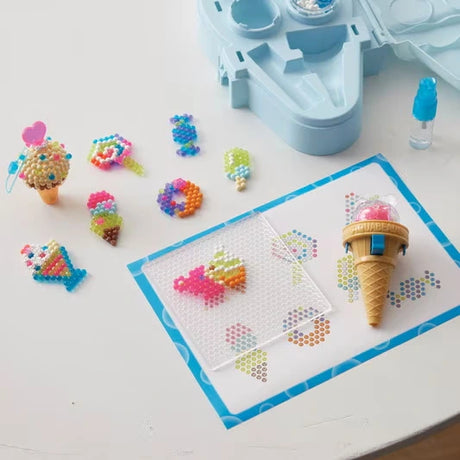 Aquabeads ice cream shaped bead pen resting on a transparent pegboard over colourful design templates, surrounded by completed sweets, ice cream cones and ice lolly bead designs.