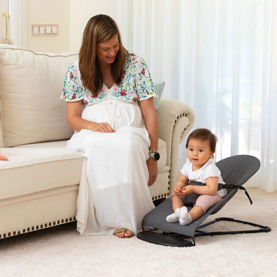 Baby seated in the grey Asalvo Paradise bouncer beside a seated woman indoors