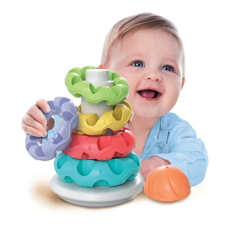 Smiling baby wearing a light blue top holding a purple textured ring beside the Baby Clementoni Stacking Rings tower, with the orange dome resting on the floor.