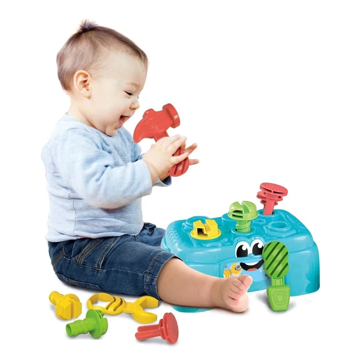 Young child sitting beside the Baby Clementoni Workbench, holding a red toy hammer, with yellow and green tools and fasteners on the floor.
