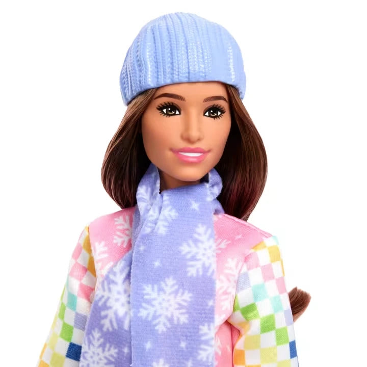 Close-up of Barbie Snowboarder Doll with brown hair, a light blue knitted beanie and matching blue snowflake scarf, wearing a pink jacket with multicoloured checked sleeves.