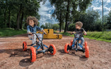 Two children seated on BERG Buzzy Nitro pedal go-karts outdoors with trees and numbered yellow blocks in the background