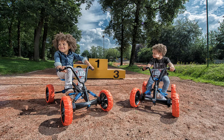 Two children seated on BERG Buzzy Nitro pedal go-karts outdoors with trees and numbered yellow blocks in the background