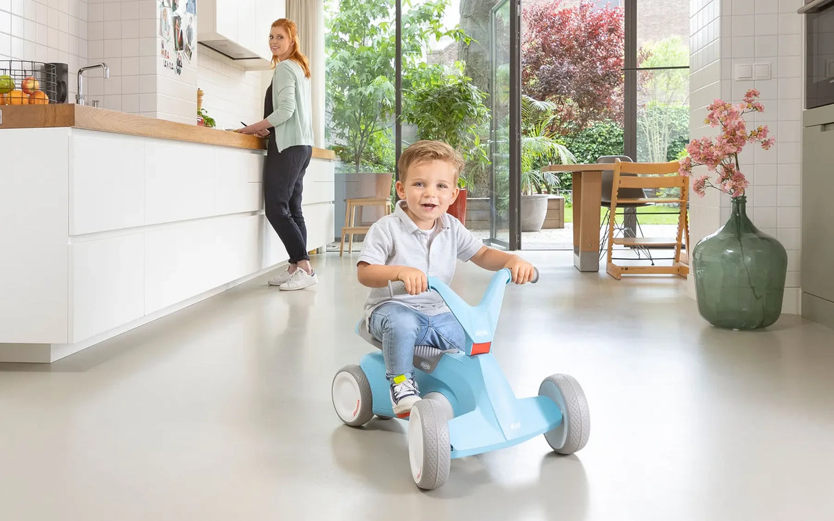 Child holding the handlebars of a blue BERG GO² ride-on indoors with an adult standing near a counter