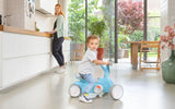Child seated on a blue BERG GO² ride-on indoors with an adult standing near a counter