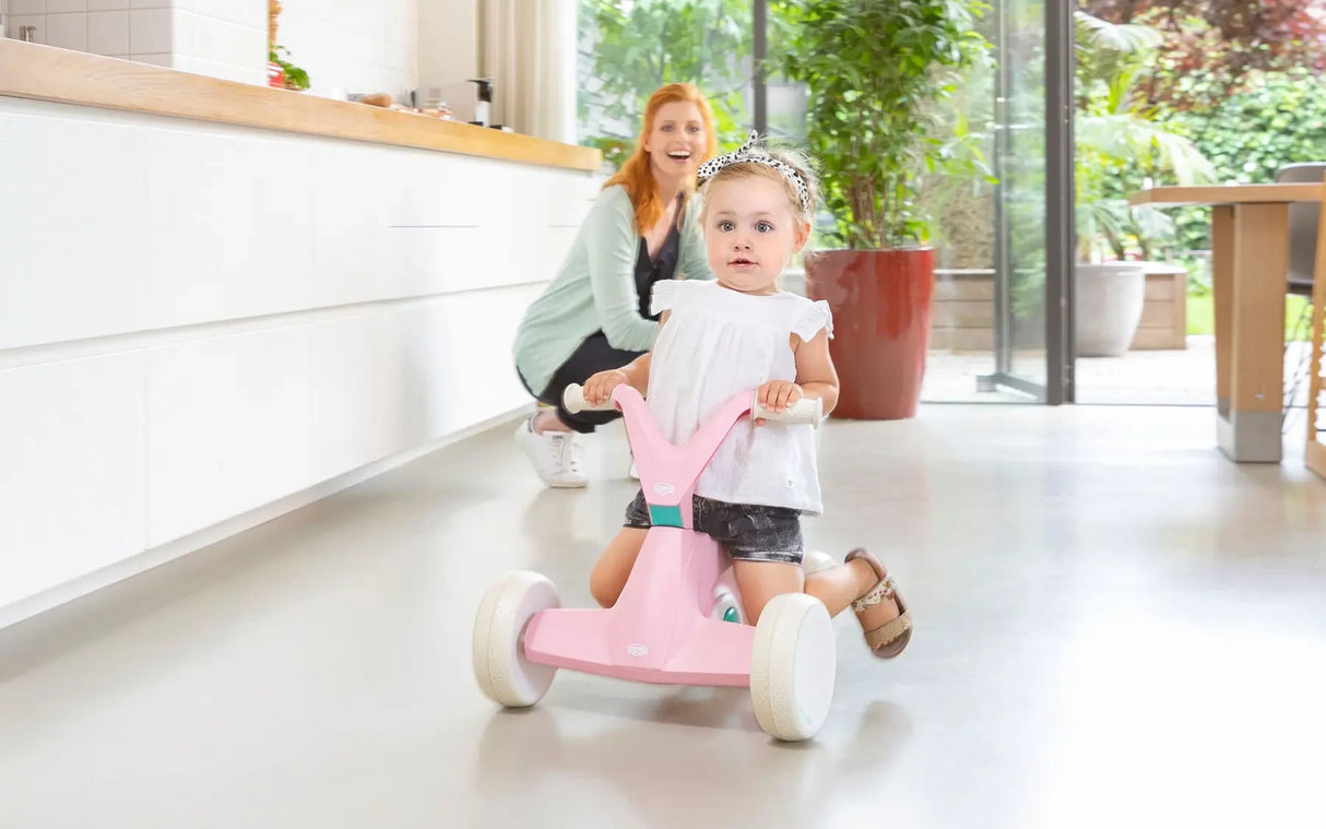 Child holding the handlebars of pink BERG GO² ride-on indoors with adult in the background