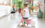 Child seated on pink BERG GO² ride-on indoors with adult crouching nearby