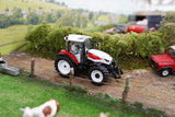 Britains Steyr 4120 Plus farm toy tractor on a model farm lane with a red trailer and cows visible