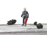 Bruder bworld Fireman with Accessories 60100 standing on a wooden surface among dark rocks, holding a red fire extinguisher and black radio while wearing protective firefighter clothing