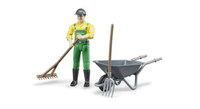 bWorld farmer figure wearing green dungarees and a yellow shirt, pushing a grey wheelbarrow carrying a brown besom broom and a grey pitchfork on a white background.