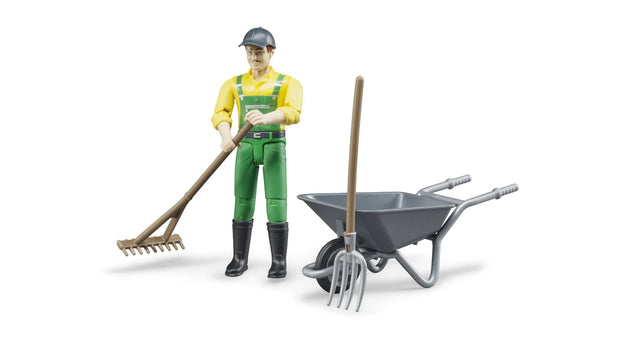 bWorld farmer figure wearing green dungarees and a yellow shirt, pushing a grey wheelbarrow carrying a brown besom broom and a grey pitchfork on a white background.