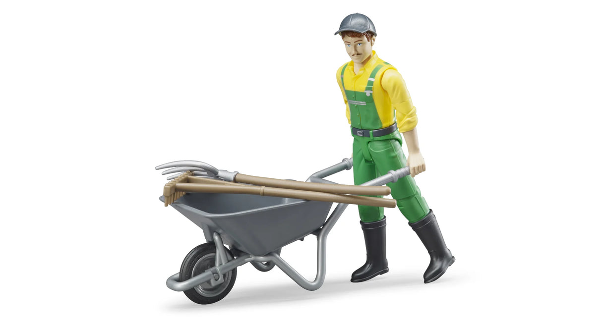 bWorld farmer figure wearing green dungarees and a yellow shirt, standing beside a grey wheelbarrow with a brown besom broom and a grey pitchfork on a white background.