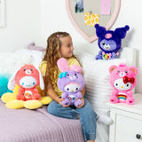 Young girl sitting on a bed holding purple My Melody as Share Bear plush, surrounded by three other Care Bears x Hello Kitty character plush toys in orange, pink and dark purple.