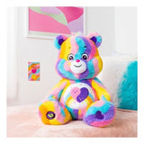 Care Bears Jumbo Plush Friends Forever Bear seated on a pink surface in a bedroom setting, showing the full bear with multicolour striped fur and double heart tummy badge