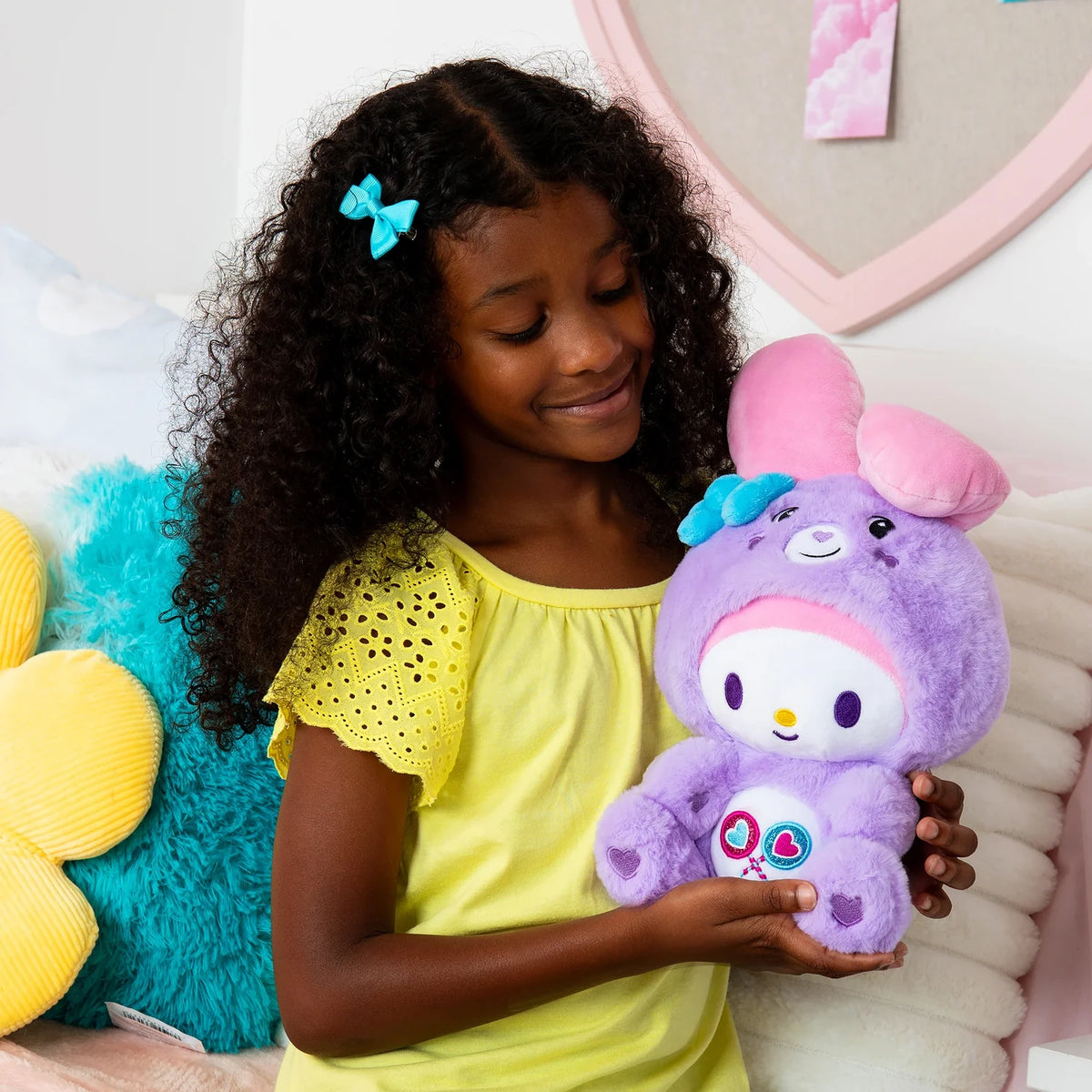 Young girl wearing a yellow top and turquoise hair bow, holding the purple My Melody as Share Bear plush with its pink ears and heart lollipop belly badge visible.
