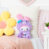 My Melody as Share Bear plush sitting on a pink bedspread in front of a white cushion, beside a large yellow flower cushion.