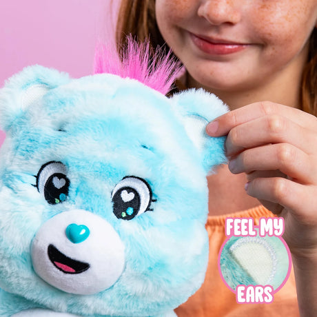 Child touching the textured ear of Care Bears Sweet Serenity Bear, showing its fluffy blue fur, pink hair tuft and white sensory fabric inside the ear.