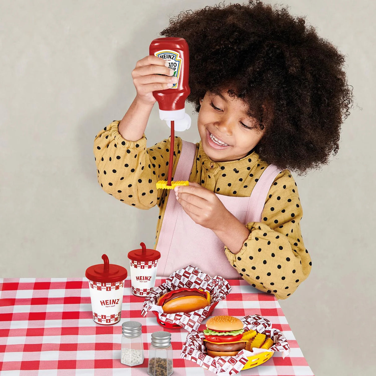 Child holding a Heinz ketchup bottle over toy diner food on a checked table