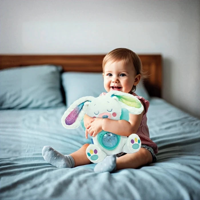 Smiling baby sitting on a bed and hugging the white Arctic Bunny plush, showing its multicoloured ears, embroidered sleeping face and turquoise projector housing.
