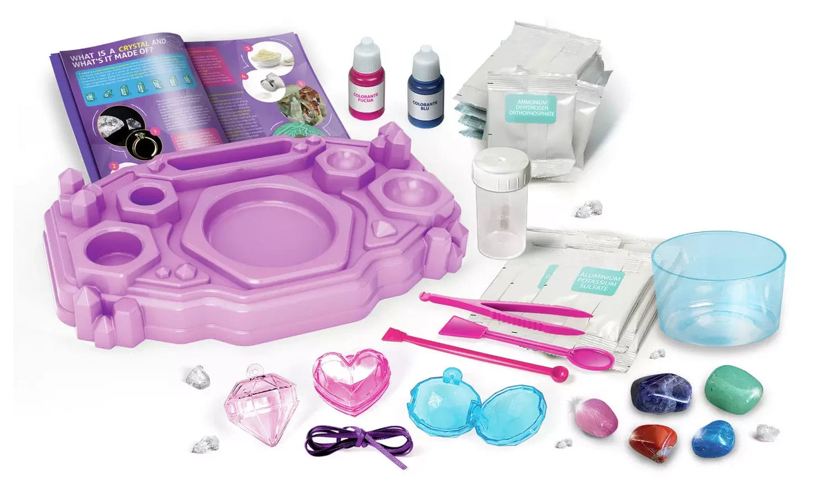 Crystal Jewellery Creator kit contents arranged on a white background, including purple workstation, instruction booklet, chemical packets, colour bottles, measuring containers, pink tools, gemstone specimens and jewellery charms.