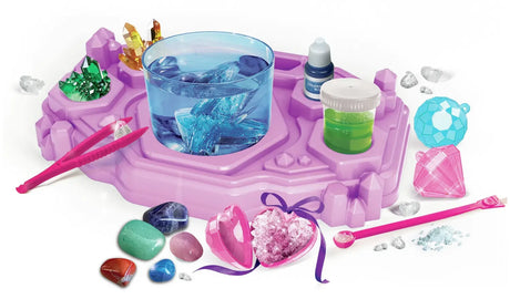 Purple Crystal Jewellery Creator workstation with a transparent blue cup containing crystals, green and yellow crystal clusters, pink tweezers, coloured stones and a pink heart shaped jewellery container.