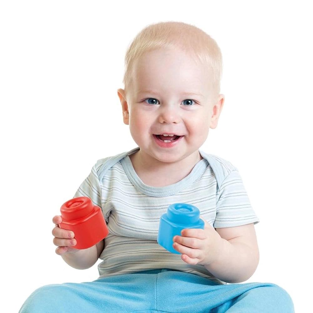 Baby sitting and holding two Clementoni Soft Clemmy blocks, one red and one blue in each hand.