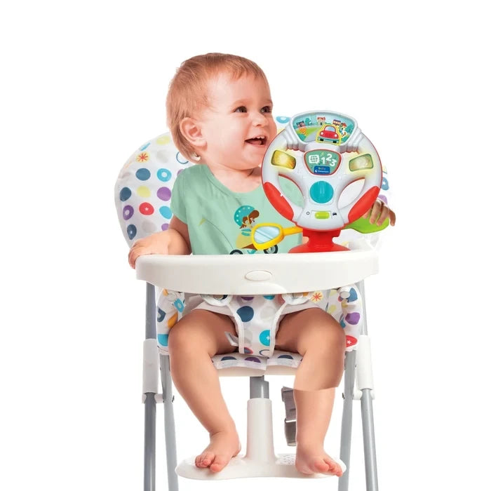 Smiling baby wearing a green T-shirt sitting in a patterned highchair with the Baby Clementoni Steering Wheel positioned on the white tray.