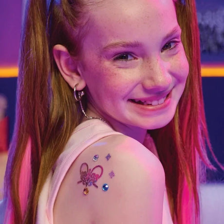 Smiling girl with long red hair showing a shimmering heart shaped temporary tattoo decorated with purple, blue and gold gems on her shoulder.