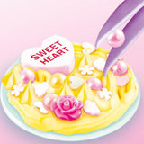 Close-up of yellow Creamy Deco cream piped onto a round blue base, decorated with a Sweet Heart charm, pink pearls, white flowers, small hearts and a pink rose, with purple tweezers positioning a decoration.