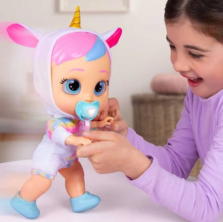 Young girl wearing a purple top holding both hands of Cry Babies My First Steps Dreamy as the doll takes a step, wearing her unicorn outfit and turquoise shoes with a dummy in her mouth.