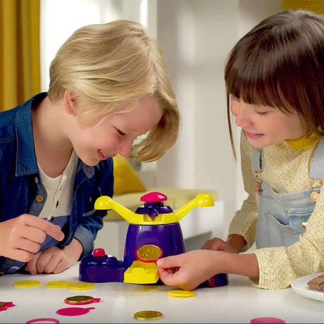Two children using the John Adams Golden Coin Maker at a table, with one child holding a finished golden coin beside the purple and yellow machine, moulds and chocolate pieces