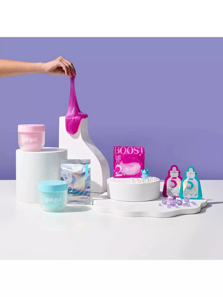 Gui Gui Deluxe Twin Pack pink shimmer slime being stretched upward from a white pedestal display, with teal and pink jars, base sachet, booster, figurine, charm beads and two sparkle sachets arranged alongside