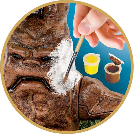 Close-up of a brown Harry Potter Mandrake figure being painted with a small brush, showing white paint on its face and two open paint pots containing yellow and brown paint.