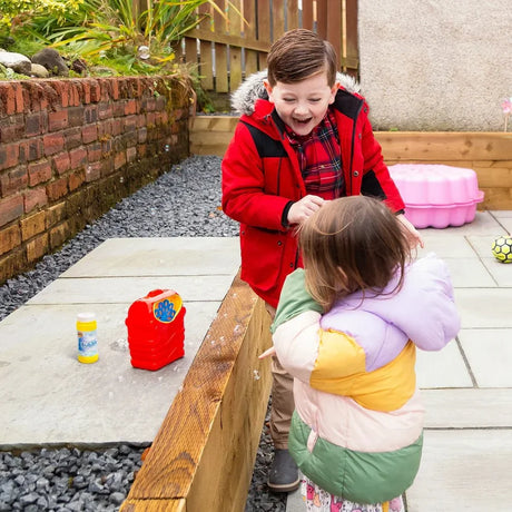 Two children playing outdoors with the HGL Bubble Machine placed on a raised garden bed beside the bubble solution bottle, with bubbles visible in the background