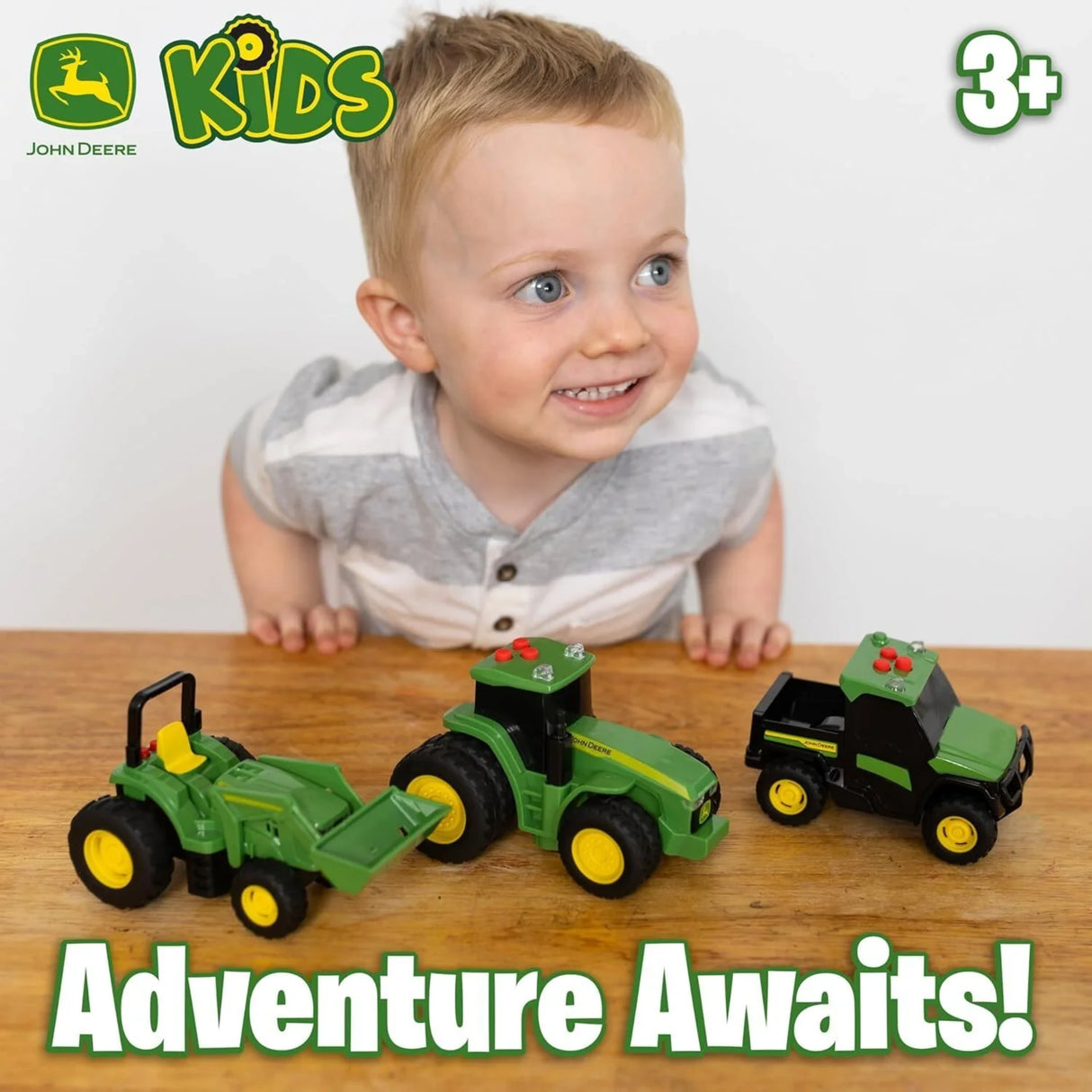 Child leaning over wooden table with three John Deere mini vehicles, John Deere KIDS logo, “3+” age mark and “Adventure Awaits!” text