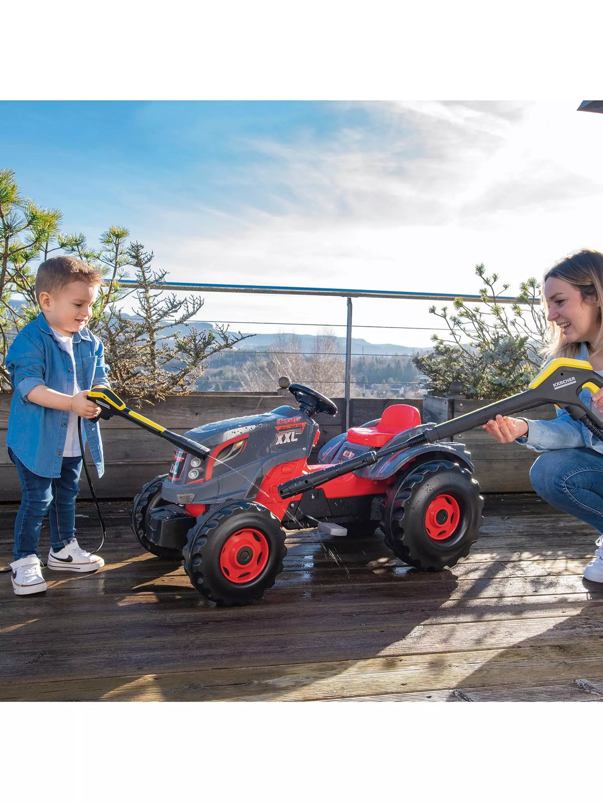 Child and woman using the Smoby Karcher High Pressure Trolley K4 to spray a red ride-on tractor outdoors
