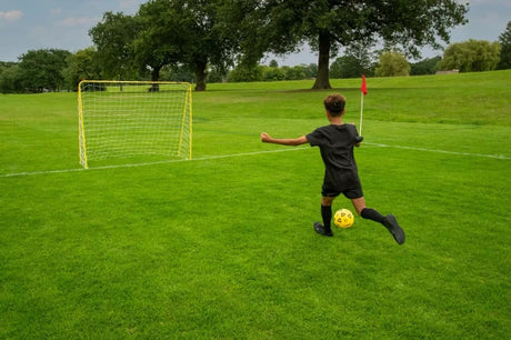 Child kicking a yellow football towards a yellow metal goal with white netting on grass 