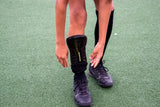 Child adjusting a black shin pad worn under a black sock on an outdoor artificial pitch