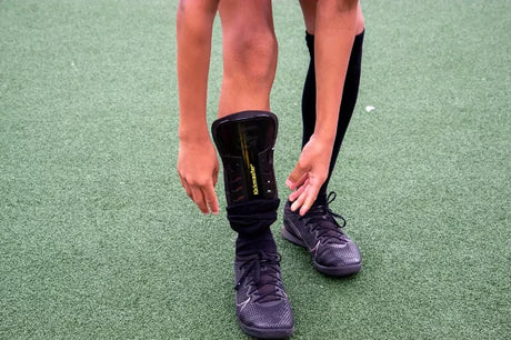 Child adjusting a black shin pad worn under a black sock on an outdoor artificial pitch