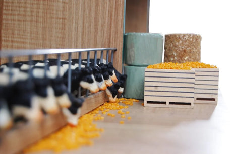 Close-up of Kids Globe Farming 1:32 farm accessory pieces including wooden crates, wrapped round bales and square hay bale alongside dairy cow figures inside a farm building