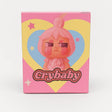 L Monsters collectible toy packaging featuring a grumpy pink cartoon character with rosy cheeks in front of a yellow and blue heart and stars, labeled ‘Crybaby’.