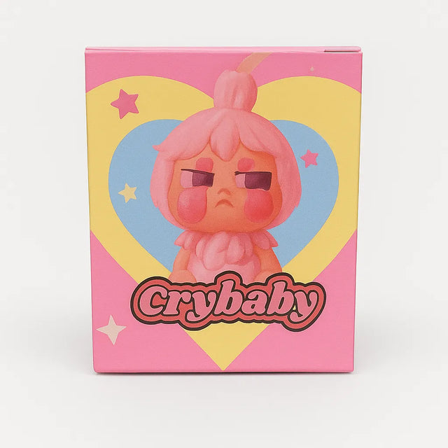 L Monsters collectible toy packaging featuring a grumpy pink cartoon character with rosy cheeks in front of a yellow and blue heart and stars, labeled ‘Crybaby’.
