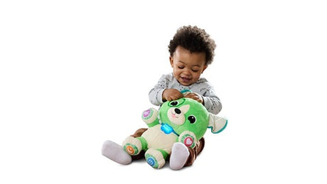 Young child sitting on the floor pressing the interactive buttons on a LeapFrog My Best Pal Scout plush puppy