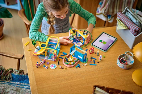 Child building LEGO Friends Heartlake City Friends Club House 42689 at a wooden table, with clubhouse sections, minidolls, loose bricks and a tablet displaying building instructions.