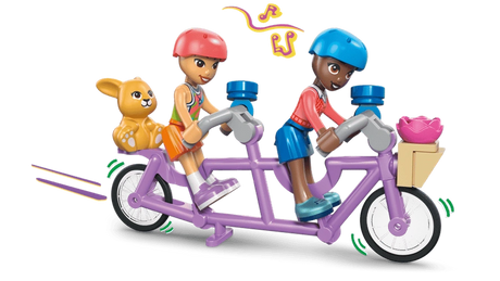 Two LEGO Friends minidolls wearing pink and blue helmets riding a purple tandem bicycle, with a brown bunny in the rear basket and pink flower in the front basket.