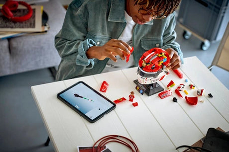 Person assembling the LEGO Scuderia Ferrari Charles Leclerc Helmet 43014 at a table with loose LEGO pieces, minifigure and tablet displaying digital building instructions.