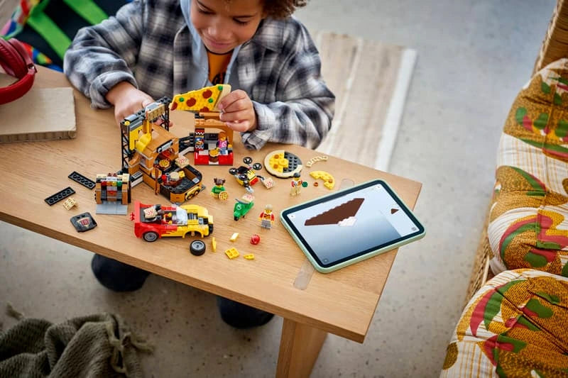 Child building a LEGO City pizza delivery set on a table with sports car, scooter, minifigures and tablet instructions