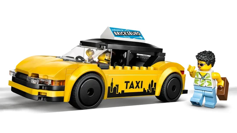 Front angled view of the LEGO City yellow taxi showing the yellow body, black roof, white roof sign, TAXI text on the side panel, and a minifigure standing next to the front right side of the car