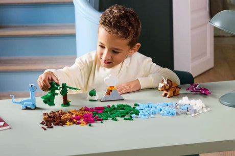 Child building LEGO Classic Creative Dinosaurs 11041 at a table with dinosaur models, volcano, palm tree and colourful bricks